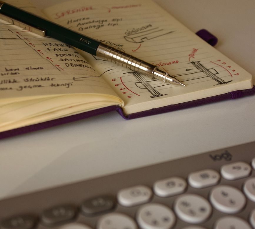 A notebook with handwritten notes and diagrams, featuring a silver pen resting on top, beside a gray keyboard.
